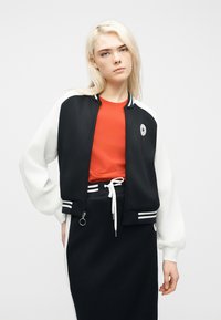 Νεαρή γυναίκα με ξανθά μαλλιά, ντυμένη με μαύρο και άσπρο μπουφάν, κόκκινο πουκάμισο και μαύρη φούστα με άσπρες ρίγες, στέκεται μπροστά σε απλό φόντο.