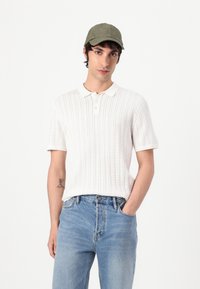 Hvid ribbet strik polo skjorte med korte ærmer og krave, kombineret med lyseblå denim jeans og en grøn kasket.