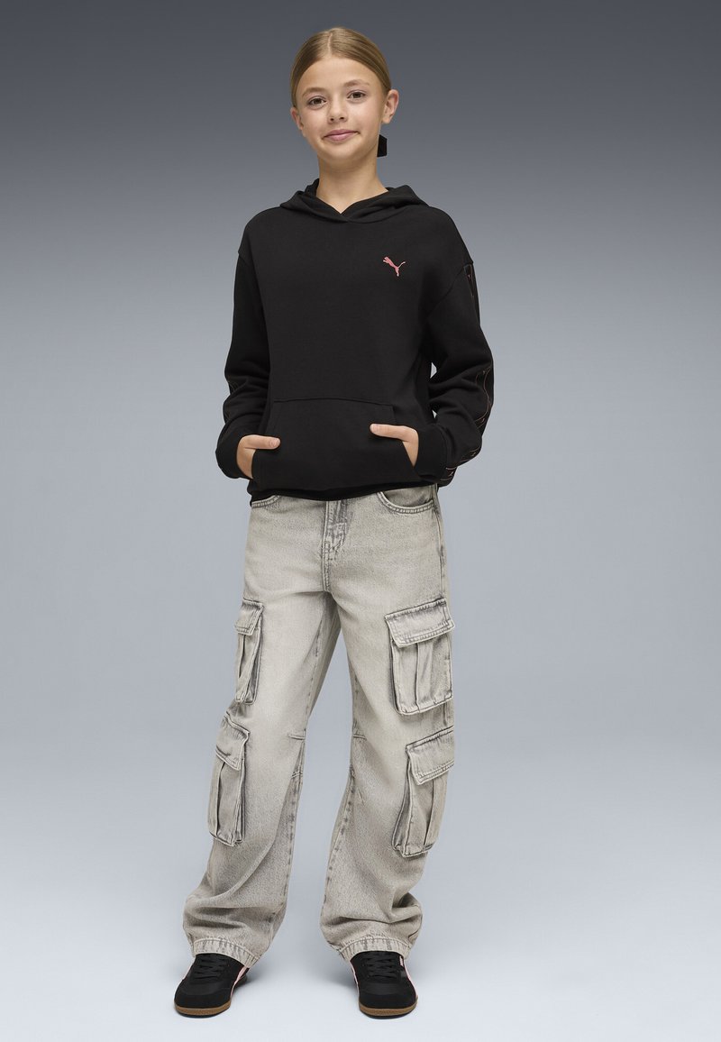 Jeune personne debout, mains dans les poches, portant un sweat à capuche noir, un pantalon cargo gris clair et des baskets noires, sur un fond gris uni.