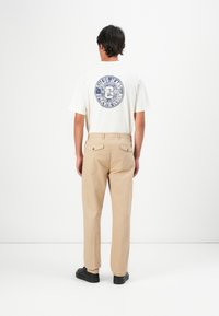 Muškarac u bež hlačama, crnim cipelama i bijelom majici s plavim kružnim logotipom "GUESS JEANS" na leđima, stoji uz bijeli zid.