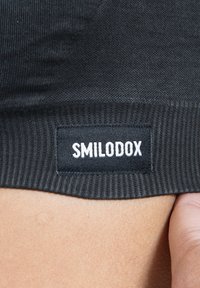 Musta kangas, jossa on ribbikuvio, ja siinä on ommeltu etiketti, jossa on valkoisella tekstillä lukemassa "SMILODOX". Läheltä nähtynä näkyy alahelman yksityiskohta.