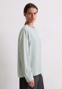 Hellgrüne Bluse aus seidig-weichem Material, lange Ärmel mit Knopfmanschetten, runder Halsausschnitt, subtile Vorderfalte, kombiniert mit dunklen Hosen.