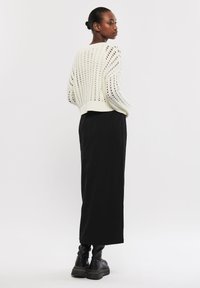 Pull en tricot ajouré crème avec une coupe cropped, associé à une longue jupe noire et des bottes noires montantes. Texture lisse, quincaillerie minimale.