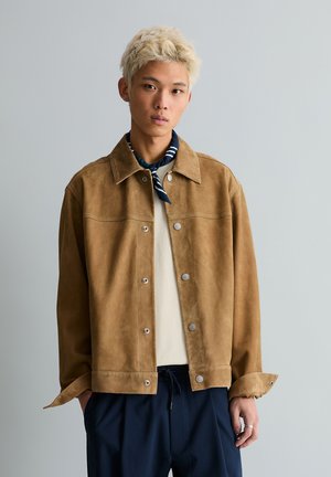 Junger Mann mit gebleichtem Haar trägt eine hellbraune Wildlederjacke, cremefarbenes Hemd, marineblaue Hose und einen marineblau gestreiften Halstuch vor grauem Hintergrund.