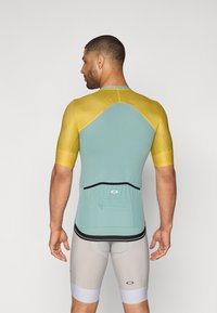 Idretts skjorte med korte ermer; mintgrønn kropp, gule skuldre, nettingtekstur, svarte detaljer og en baklomme. Grå shorts med hvit kant.