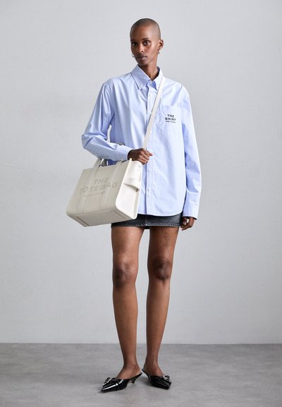 Geantă tote din piele albă cu inscripția "THE TOTE BAG" embosată, purtată peste o cămașă cu nasturi de culoare albastru deschis și o fustă mini din denim, asociată cu mule negre cu toc.
