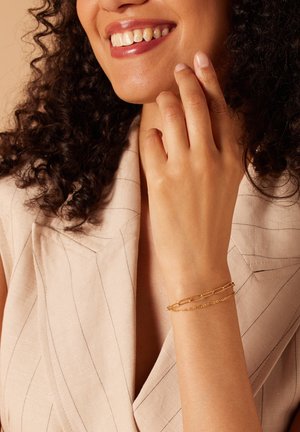 Gouden kettingarmband met aangesloten segmenten en een delicate ketting, gedragen om een pols met een lichte huidskleur, tegen een beige achtergrond.
