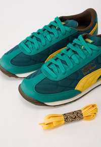 Teal a žlté Puma Easy Rider tenisky majú kombináciu štruktúrovanej tkaniny a hladkej kože, s hrubou hnedou gumovou podrážkou. Obsahujú žlté šnúrky.
