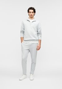 Ανοιχτό γκρι φούτερ με φερμουάρ και διακριτικό λογότυπο, σε συνδυασμό με αντίστοιχο κολάν με στενή γραμμή. Φοράται με άσπρα αθλητικά παπούτσια, πάνω σε απλό φόντο.