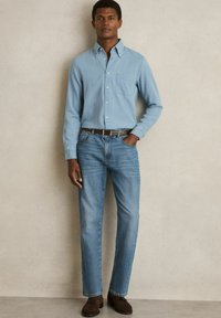 Lichtblauw overhemd met knopen en een borstzak, gecombineerd met medium-wash rechte jeans en donkere suède schoenen, voor een neutrale achtergrond.