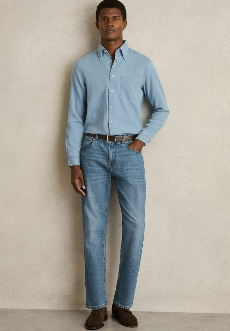 Lichtblauw overhemd met knopen en een borstzak, gecombineerd met medium-wash rechte jeans en donkere suède schoenen, voor een neutrale achtergrond.