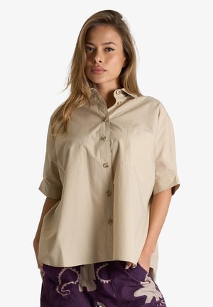 Donna con i capelli lunghi che indossa una camicia beige a maniche corte con bottoni e pantaloni viola con fantasie, in piedi su uno sfondo bianco.