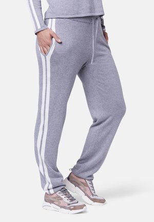 Personne portant un pantalon de survêtement gris avec des bandes blanches sur les côtés, mains dans les poches, associée à des baskets épaisses roses et grises sur fond blanc.