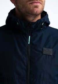 Námořnická vodotěsná bunda s vysokým límcem, předním zipem a světle modrými detaily. Obsahuje texturovanou nášivku loga na hrudi.