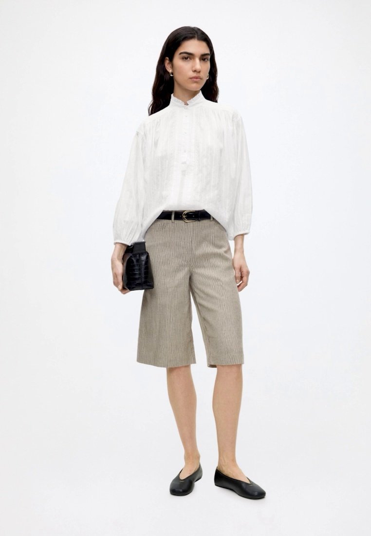 Mujer con blusa blanca de cuello alto, pantalones cortos beige a rayas hasta la rodilla, cinturón negro, bailarinas negras y sosteniendo un bolso de mano negro.