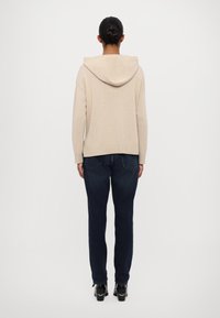 Personne aux cheveux foncés attachés en chignon, portant un pull à capuche beige, un jean bleu foncé et des bottines noires, debout de dos.