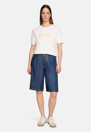 Woman wearing a white short-sleeve top with "smile" text, loose blue denim knee-length shorts, and beige-white sneakers, standing against white background.