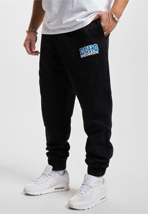Person iført sorte Ecko Unlimited joggingbukser og hvide sneakers med hænderne afslappet, den ene i lommen, mod en ensfarvet baggrund.