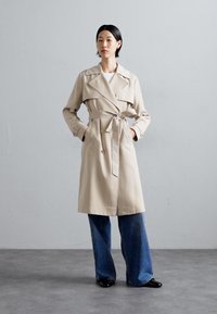 Beige trench coat with a belted waist, oversized collar, and double-breasted buttons, paired with wide-leg denim jeans and black shoes.