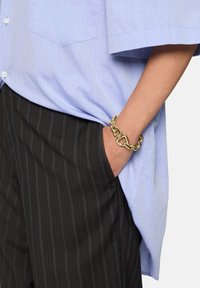 Camicia azzurro chiaro a maniche corte con taschino, abbinata a pantaloni neri a righe sottili. Al polso è indossato un bracciale in oro spesso.