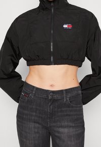 Černá zkrácená větrovka se zipem, vysokým límcem a nášivkou Tommy Jeans; v kombinaci s tmavě šedými džíny.