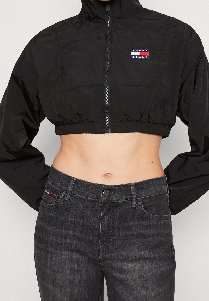 Černá zkrácená větrovka se zipem, vysokým límcem a nášivkou Tommy Jeans; v kombinaci s tmavě šedými džíny.