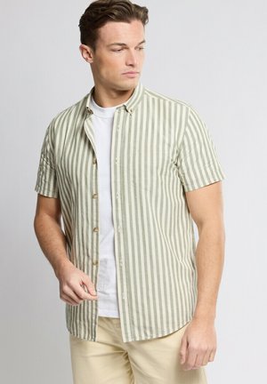 Jeune homme portant une chemise à manches courtes beige et blanche à rayures verticales par-dessus un t-shirt blanc, pantalon beige, regardant sur le côté sur un fond uni.