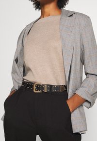 Šedý kostkovaný blejzr přes béžový svetr, spárovaný s černými kalhotami a černým opaskem s cvočky, který má zlatou sponu.