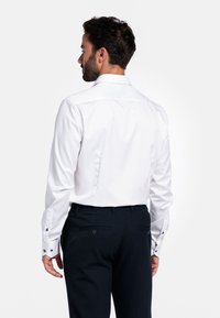 Witte katoenen overhemden met een standaardkraag en met knopen afgewerkte manchet, gecombineerd met donkere broek met achterzakken en een knopenaccent.