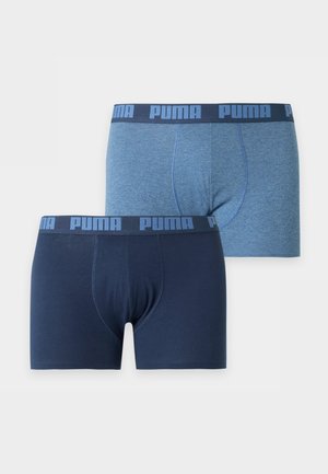 Jeune homme à la peau foncée debout pieds nus en boxer bleu Puma sur un fond blanc uni, une main derrière le dos.