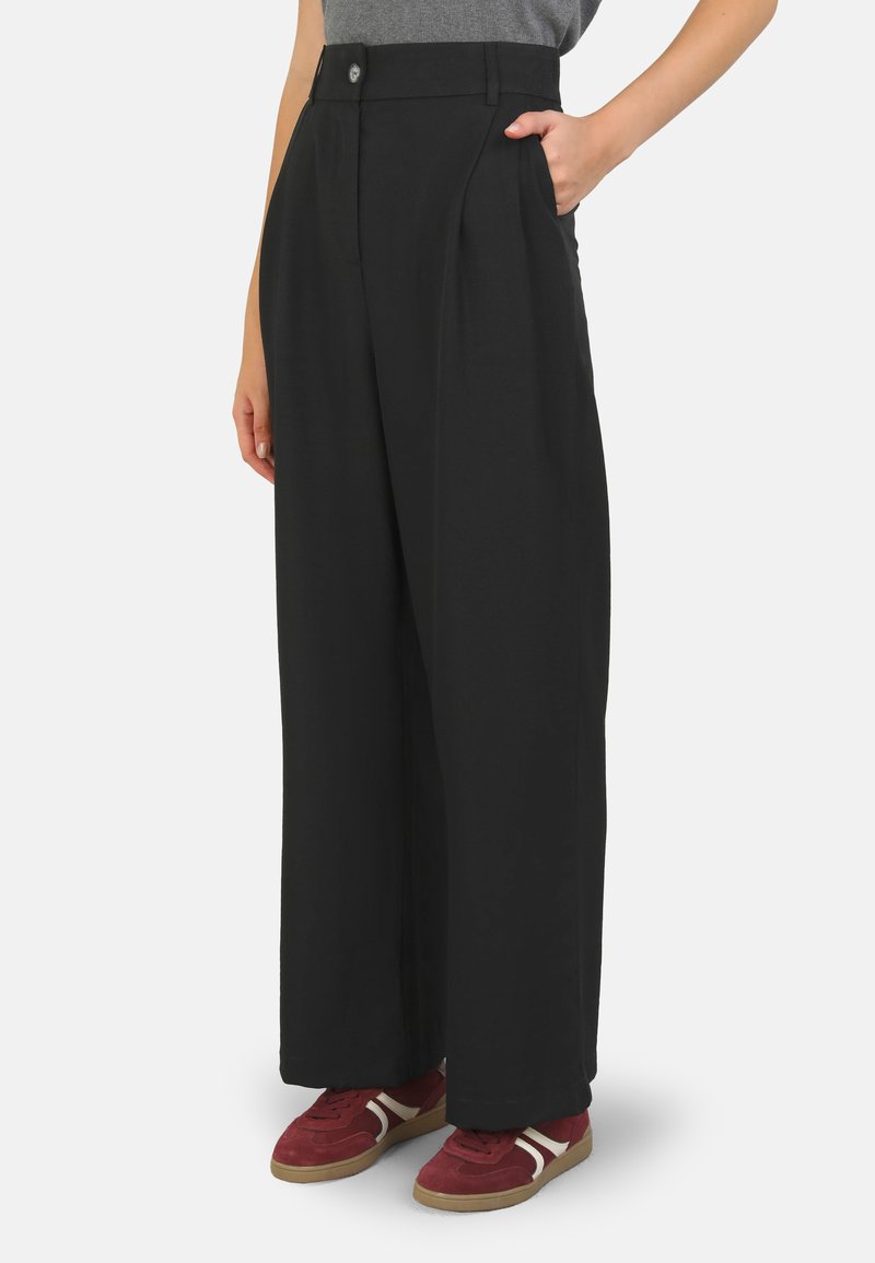 Black wide-leg trousers with pleats, a front button closure, and side pockets. Made of smooth fabric, paired with red sneakers.