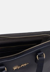 Námořnická kožená kabelka se zlatým zipem a logem Tommy Hilfiger, ukazující otevřený interiér s více přihrádkami a vnitřní kapsou na zip.