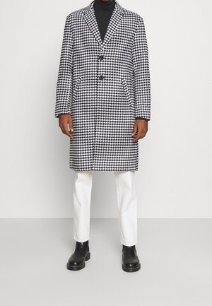Personne portant un manteau à motif pied-de-poule noir et blanc par-dessus un col roulé noir, un pantalon blanc et des bottes noires, debout devant un fond uni.