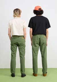 Two people stand back to back. One wears a cream short-sleeve shirt and green pants, the other a black shirt and green pants, with a cap.