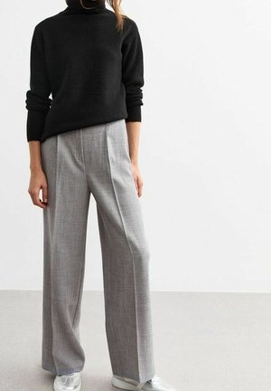 Person iført en sort langærmet sweater, lysegrå bukser med vide ben og forplissering foran samt hvide sneakers, stående foran en ensfarvet hvid baggrund.