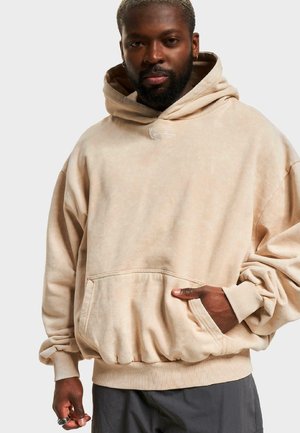 Beige oversized hoodie with a front pocket, ribbed cuffs, and hem. Features a small embroidered logo on the chest. Soft, textured fabric.