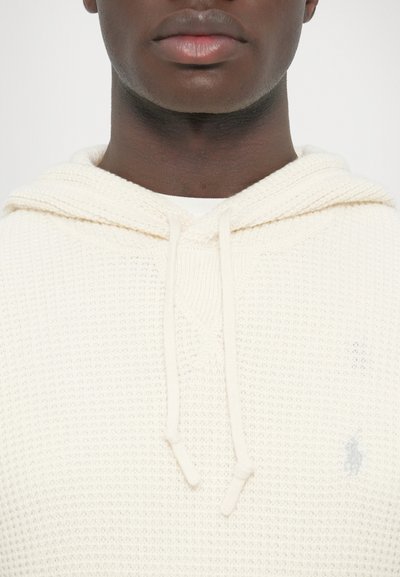 Krémovo sfarbená pletená mikina s kapucňou so štruktúrovaným vzorom, sťahovacími šnúrkami a mal�ým logom na ľavej strane. Pohľad spredu.