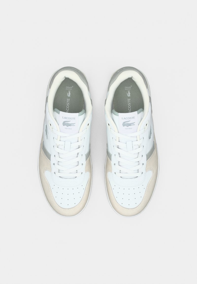 Sneakers Lacoste con tomaia in pelle bianca, dettagli grigi e tallone in pelle beige. Presentano fori di aerazione e lacci bianchi.