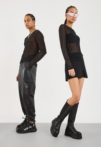 Schwarzes Netzoberteil mit langen Ärmeln, schwarze Cargo-Hosen aus Leder und schwarze Plateaustiefel an einem männlichen Modell; weibliches Modell in einem schwarzen enganliegenden Oberteil und einem Minirock.