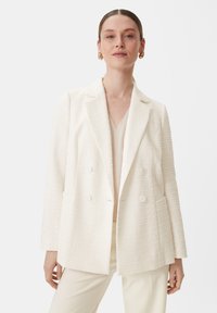 White textured cotton blend blazer, double-breasted with flat buttons, notched lapels, and side pockets. Worn over a light top.