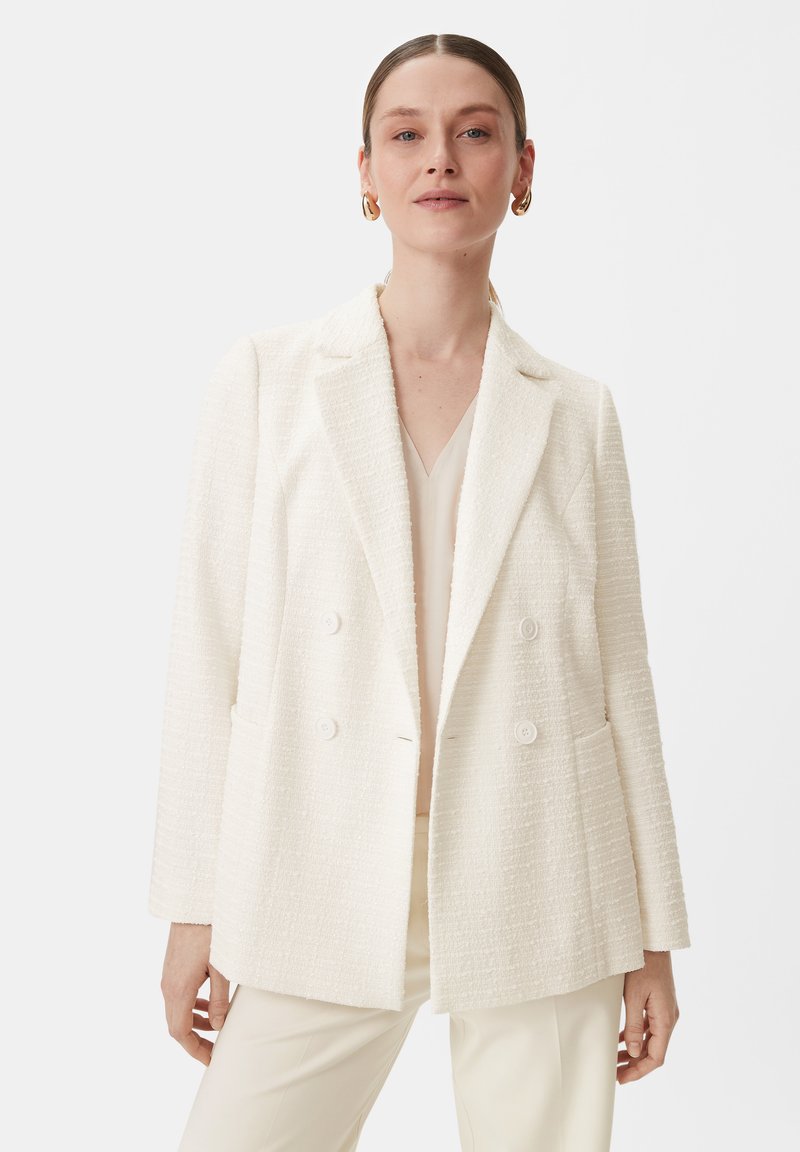 White textured cotton blend blazer, double-breasted with flat buttons, notched lapels, and side pockets. Worn over a light top.