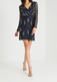 Tmavě modré šaty s dlouhým rukávem, výstřihem do V a průhledným overalem, s vlnitým vzorem a sešívanými akcenty. Délka po kolena s přiléhavým živůtkem.