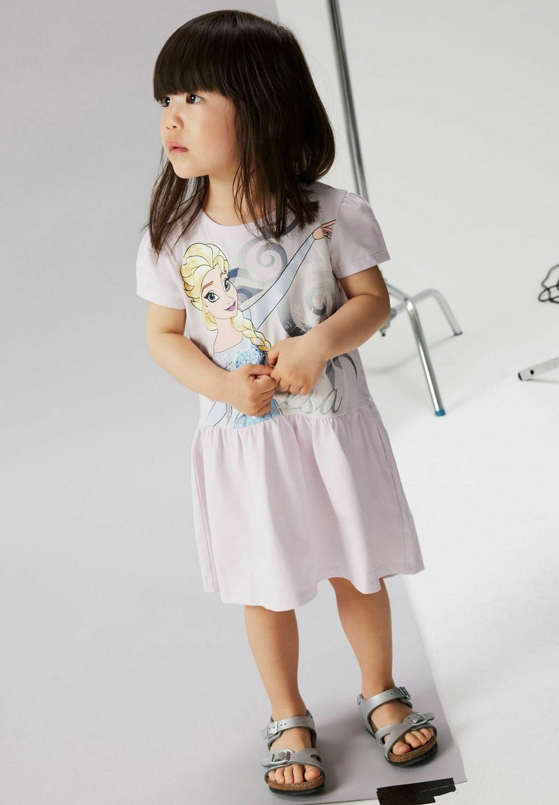 Ragazza giovane che indossa un vestito rosa chiaro con stampa di Elsa di Frozen, in piedi a piedi nudi su un pavimento bianco con attrezzature da studio sullo sfondo.