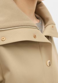 Chaqueta beige con un gran cuello, que presenta herrajes dorados, cierre de cremallera y botones a presión. El material parece suave y ligeramente texturizado.