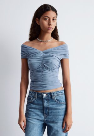 Light blue off-the-shoulder top featuring gathered fabric at the bust and waist, paired with high-waisted light blue jeans.