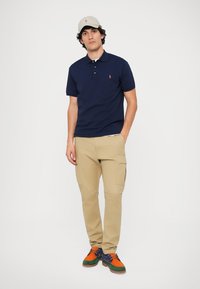 Temno modra polo majica, kaki cargo hlače in barvita športna obutev. Dodatki vključujejo svetlo kapo. Sproščena oblačila s kontrastnimi barvami.