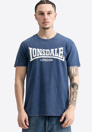 Mladý muž s vrkočmi, oblečený v modré tričko Lonsdale London, ukazuje detailné tetovania na pravom predlaktí.