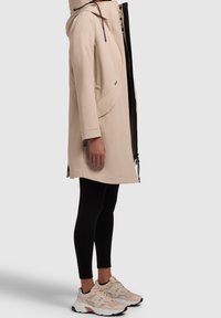 Veste beige résistante à l'eau avec capuche, poches latérales et fermeture éclair frontale. Associée à des leggings noirs et des baskets légères.