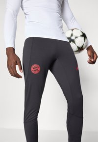 Sorte treningsbukser med teksturert stoff, med en rød FC Bayern München-logo på låret og broderte detaljer langs sømmene.