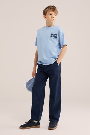 Dreng iført lyseblå grafisk t-shirt, mørkeblå bukser og sorte sneakers, holder en lyseblå kasket i den ene hånd og kigger til siden.